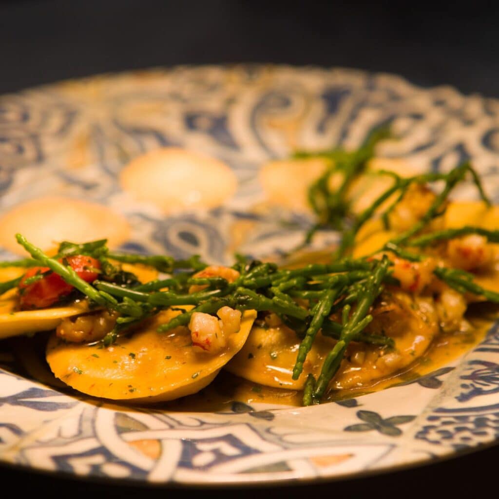 First course of fish with ravioli and sea asparagus on decorated plate.